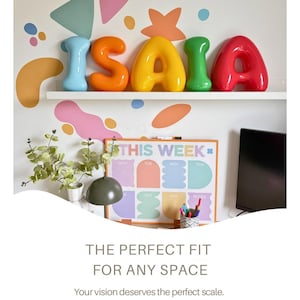 May include: Five glossy, rounded, three-dimensional letters spelling ISAIA in light blue, orange, yellow, green, and red. The letters are arranged on a white shelf against a wall decorated with colorful abstract shapes and a weekly planner board.