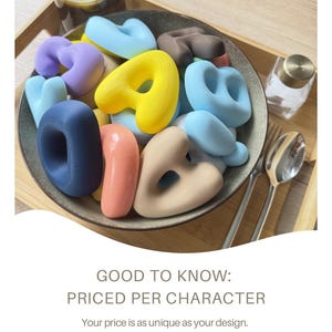 May include: A collection of smooth, matte-finish alphabet letters in various pastel and bold colors, including yellow, blue, pink, purple, and dark brown, contained within a dark gray bowl. The text GOOD TO KNOW: PRICED PER CHARACTER is visible.