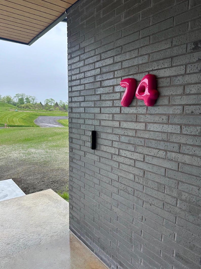 May include: A set of glossy, bright pink house numbers displaying the digits 74, mounted on a dark gray brick wall. The numbers have a rounded, three-dimensional appearance and are positioned to the right of a vertical black rectangular device.
