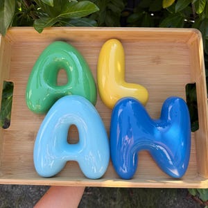 May include: Four glossy, rounded, three-dimensional letters spelling the name ALAN. The letter A is green, the letter L is yellow, the second A is light blue, and the letter N is dark blue. These smooth, balloon-style letters are arranged inside a wooden tray.
