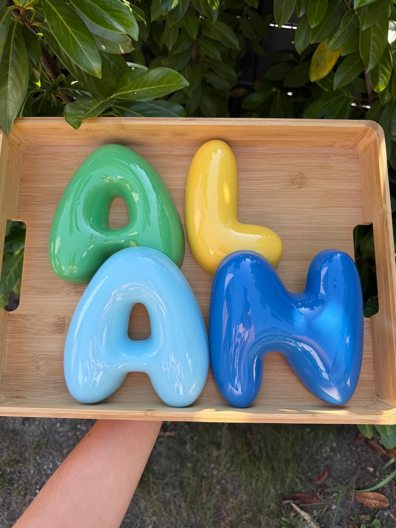 May include: Four glossy, rounded, three-dimensional letters spelling the name ALAN. The letter A is green, the letter L is yellow, the second A is light blue, and the letter N is dark blue. These smooth, balloon-style letters are arranged inside a wooden tray.