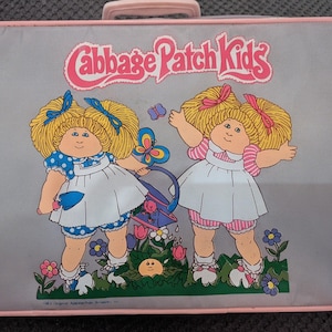 May include: A rectangular gray suitcase with a pink handle and trim featuring the Cabbage Patch Kids logo in pink text. The graphic shows two blonde dolls in white dresses with blue and pink accents, holding a watering can in a garden setting.