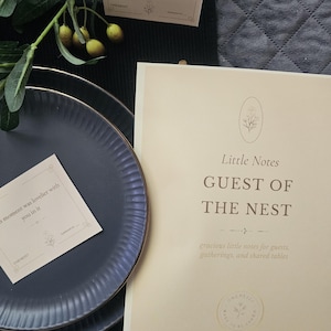 May include: A still-life arrangement with a dark blue plate with a gold rim, a card that reads "this moment was lovelier with you in it", and a larger card with the text "Little Notes GUEST OF THE NEST". Greenery with yellow berries is in the background.
