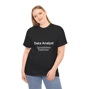 May include: Black t-shirt featuring the white text "Data Analyst Spreadsheet Detective". The shirt is worn by a person with blonde hair and blue jeans.