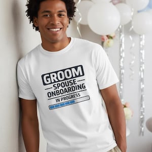 Może przedstawiać: Biała koszulka z napisem "GROOM - SPOUSE - ONBOARDING IN PROGRESS" w kolorze czarnym i niebieskim. Koszulka jest noszona przez uśmiechniętą osobę. W tle widoczne są balony i dekoracje.
