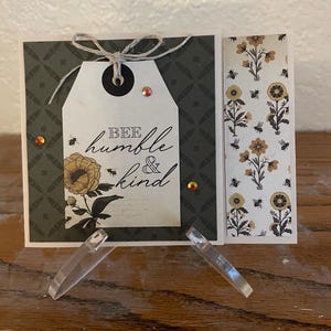 Puede incluir: Tarjeta de felicitaci&oacute;n hecha a mano con una etiqueta que dice "Bee humble & kind". La tarjeta presenta un dise&ntilde;o floral con abejas y un fondo estampado. Adornada con hilo y pedrer&iacute;a.