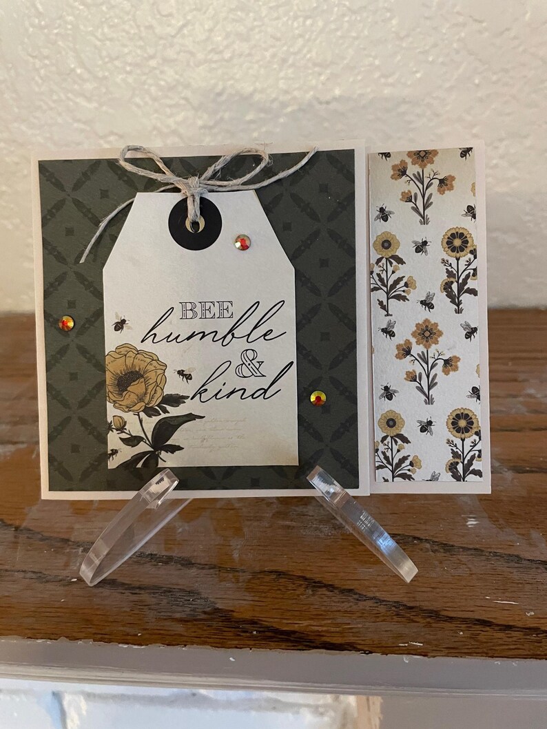 Puede incluir: Tarjeta de felicitaci&oacute;n hecha a mano con una etiqueta que dice "Bee humble & kind". La tarjeta presenta un dise&ntilde;o floral con abejas y un fondo estampado. Adornada con hilo y pedrer&iacute;a.