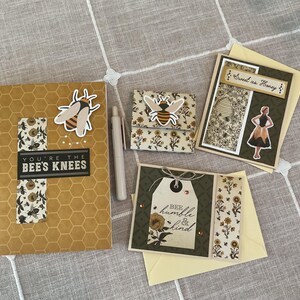 Puede incluir: Una colecci&oacute;n de art&iacute;culos de papeler&iacute;a con tem&aacute;tica de abejas, que incluye un cuaderno con el texto "You're the Bee's Knees", una cartera peque&ntilde;a y tarjetas de felicitaci&oacute;n. Los art&iacute;culos presentan dise&ntilde;os de abejas y flores en amarillo, negro y blanco.