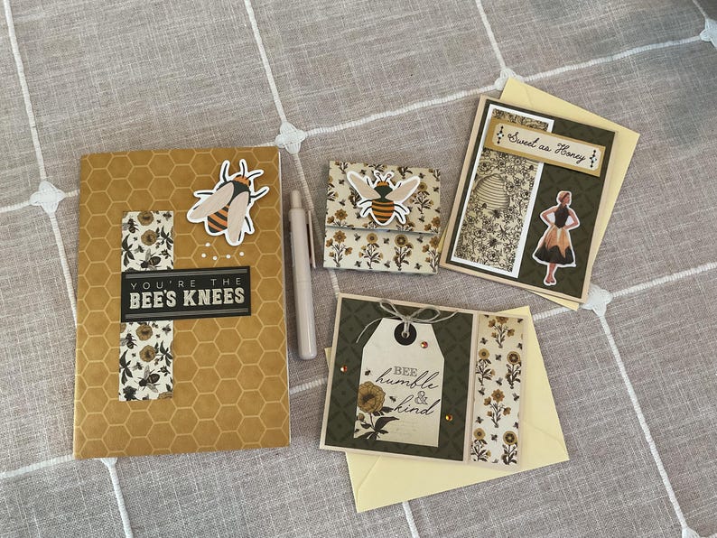 Puede incluir: Una colecci&oacute;n de art&iacute;culos de papeler&iacute;a con tem&aacute;tica de abejas, que incluye un cuaderno con el texto "You're the Bee's Knees", una cartera peque&ntilde;a y tarjetas de felicitaci&oacute;n. Los art&iacute;culos presentan dise&ntilde;os de abejas y flores en amarillo, negro y blanco.