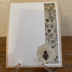 Puede incluir: Una tarjeta de felicitaci&oacute;n beige con interior blanco, exhibida en un soporte acr&iacute;lico transparente. La tarjeta presenta un dise&ntilde;o decorativo con tem&aacute;tica de abejas, que incluye una ilustraci&oacute;n de abeja y un borde estampado con abejas y elementos florales.