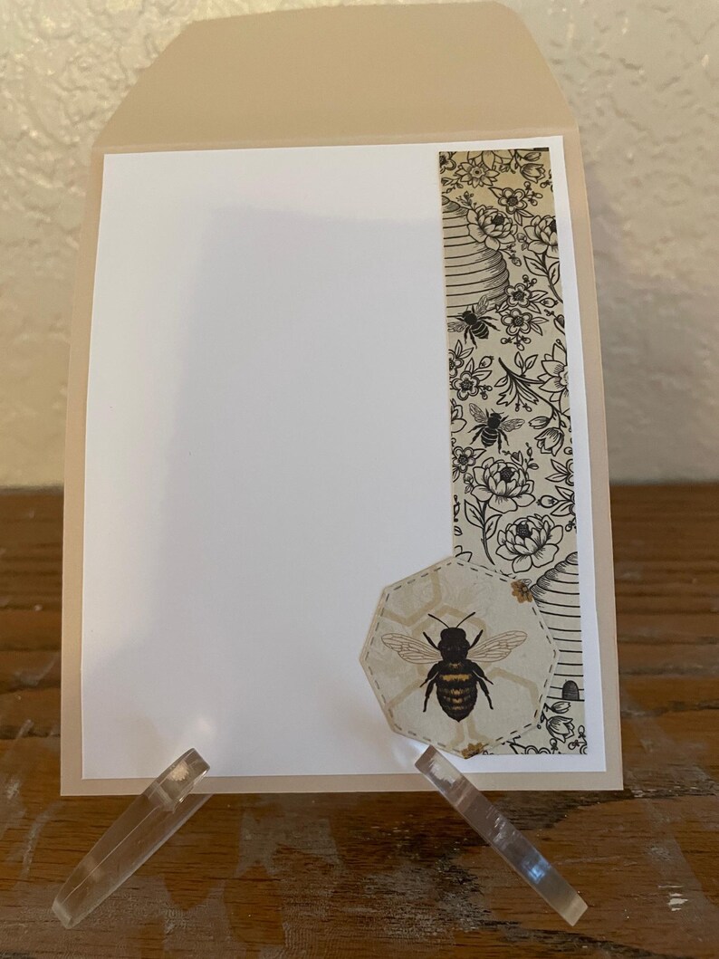 Puede incluir: Una tarjeta de felicitaci&oacute;n beige con interior blanco, exhibida en un soporte acr&iacute;lico transparente. La tarjeta presenta un dise&ntilde;o decorativo con tem&aacute;tica de abejas, que incluye una ilustraci&oacute;n de abeja y un borde estampado con abejas y elementos florales.