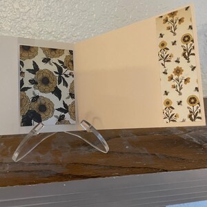 Puede incluir: Una tarjeta de felicitaci&oacute;n abierta con un dise&ntilde;o floral y de abejas. La tarjeta presenta un interior color crema y patrones florales decorativos en amarillo, negro y blanco. Se apoya en un soporte de pl&aacute;stico transparente sobre una superficie de madera.