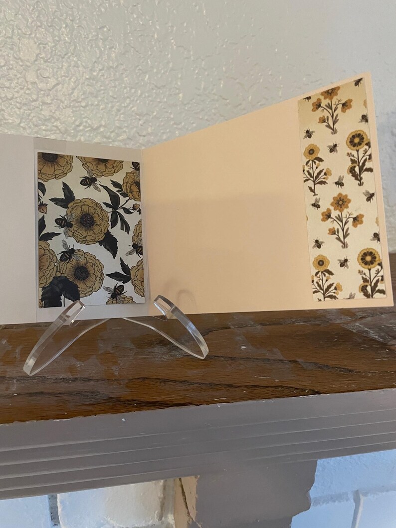 Puede incluir: Una tarjeta de felicitaci&oacute;n abierta con un dise&ntilde;o floral y de abejas. La tarjeta presenta un interior color crema y patrones florales decorativos en amarillo, negro y blanco. Se apoya en un soporte de pl&aacute;stico transparente sobre una superficie de madera.