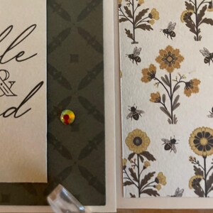 Puede incluir: Primer plano de una tarjeta de felicitaci&oacute;n hecha a mano con un patr&oacute;n floral y de abejas. La tarjeta presenta una etiqueta con escritura elegante y una gema decorativa. El fondo incluye un dise&ntilde;o geom&eacute;trico verde oscuro.