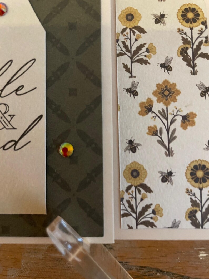 Puede incluir: Primer plano de una tarjeta de felicitaci&oacute;n hecha a mano con un patr&oacute;n floral y de abejas. La tarjeta presenta una etiqueta con escritura elegante y una gema decorativa. El fondo incluye un dise&ntilde;o geom&eacute;trico verde oscuro.