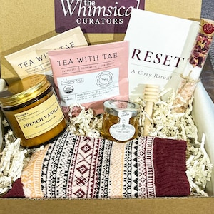 May include: A gift box filled with self-care items. Includes a jar of French vanilla candle, tea packets, a book titled "RESET", a honey jar, a wooden honey dipper, a tube of bath salts, and a pair of patterned socks.