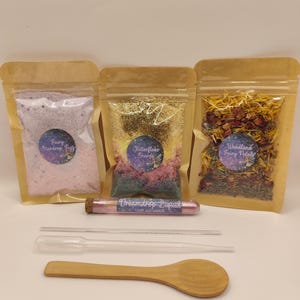 May include: A fairy-themed craft kit featuring three sealed pouches with pink, gold, and floral contents. Also included is a glass vial of "Dreamdrop Liquid", two pipettes, and a wooden spoon for mixing.