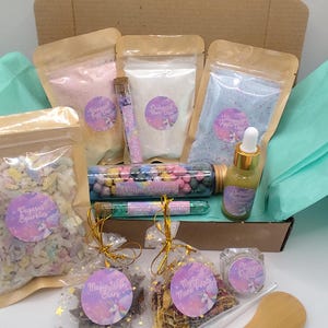 May include: A selection of colourful items presented in an open cardboard box. The box contains pouches of pink, white, and blue powder, a glass tube of sweets, and a small bottle of yellow liquid. There are also bags of confetti and dried petals.