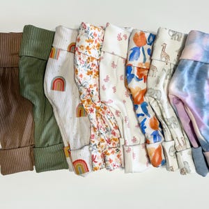 May include: A collection of baby pants in various colors and patterns. The pants include solid brown and green corduroy, white with rainbow and floral prints, and a tie-dye design. The cuffs are folded over.