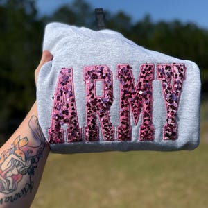 May include: A light gray sweatshirt featuring the word ARMY in large, block-style letters. The letters are filled with pink and dark purple sequins, creating a textured, shimmering appearance against the plain, heathered gray fabric of the garment.