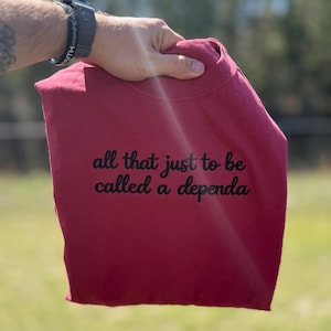 Op de afbeelding: Een bordeauxrode sweatshirt met de zwarte tekst "all that just to be called a dependa". De sweatshirt wordt door een hand omhoog gehouden, met een vage groene achtergrond.