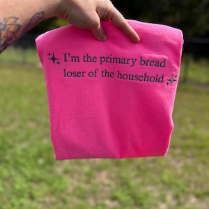 May include: A bright neon pink sweatshirt featuring dark grey embroidered text that reads, I'm the primary bread loser of the household, flanked by small four-pointed star icons on the left and right sides of the two-line phrase.