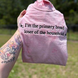 May include: A light pink crew neck t-shirt with a tie-dye effect featuring black embroidered text that reads, I'm the primary bread loser of the household, flanked by two small black star icons on the left and right sides of the lettering.
