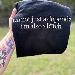Op de afbeelding: Zwart sweatshirt met witte tekst: "I'm not just a dependa, I'm also a b*tch." Het kledingstuk is opgevouwen, waarbij de boodschap centraal staat. De achtergrond is wazig, met groen gras zichtbaar.