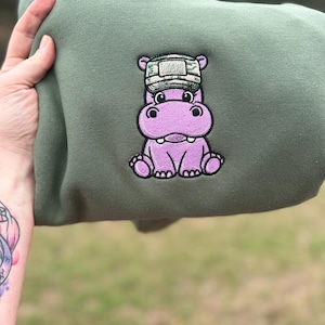 Op de afbeelding: Olijfgroene hoodie met een geborduurde paarse nijlpaardpatch met een camouflage hoed. Het geborduurde nijlpaard heeft een vriendelijke uitdrukking. De hoodie wordt door een hand omhoog gehouden, met een vage groene achtergrond.