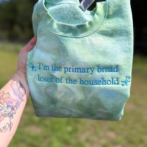 May include: A light green tie-dye crewneck sweatshirt featuring the embroidered blue text "I'm the primary bread loser of the household" flanked by small star icons. The garment has a ribbed collar and a soft interior lining visible at the neckline.