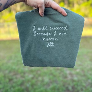 Puede incluir: Una camiseta de color verde oliva oscuro con texto bordado en blanco que dice "I will succeed because I am insane", seguido de un pequeño icono de flor de línea simple centrado debajo del texto en la parte frontal de la prenda.