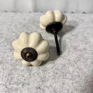 May include: Two off-white ceramic cabinet knobs shaped like flowers with a crackled glaze finish. Each knob features a dark bronze-colored metal base and a central circular metal accent with a ridged edge, attached to a threaded metal bolt.