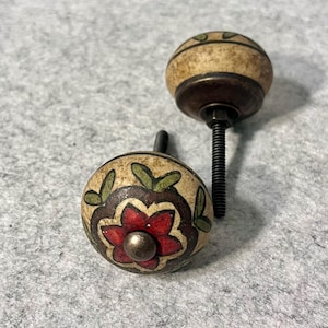 May include: Two round ceramic cabinet knobs with a cream-colored base. The front knob features a hand-painted red flower with green leaves and a dark brown outline. The back knob shows a dark metal base with a threaded bolt and a striped pattern.