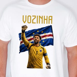 May include: A white short-sleeved t-shirt featuring a graphic print of a soccer goalkeeper in a yellow jersey with the number one, raising a fist in front of the Cape Verde flag. The text VOZINHA is printed in gold above the graphic illustration.