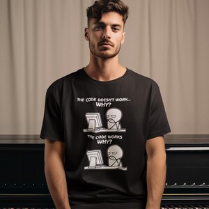 May include: A black short-sleeved t-shirt featuring a white graphic print of a stick figure at a computer. The text reads: THE CODE DOESN'T WORK... WHY? followed by THE CODE WORKS WHY? The design depicts a frustrated and then confused character.