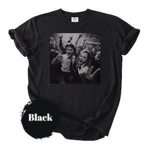 May include: A black short-sleeve t-shirt featuring a square graphic print of a crowd of people in white shirts and ties, with several individuals making rock-and-roll hand gestures. A circular black icon with the white text Black is included.