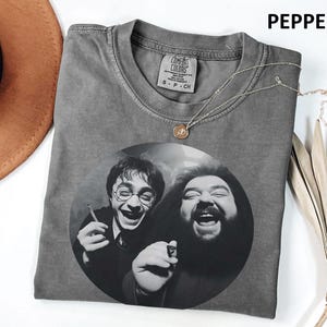 A gray t-shirt with a black and white image of two men, one with glasses and the other with a beard, smiling and holding a cigarette. The t-shirt is placed on a white surface, with a brown hat and some dried leaves nearby.