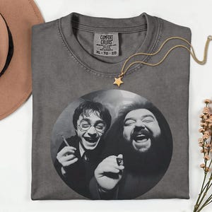 a gray t-shirt with a black and white photo of two men, one with glasses and the other with a beard, both smiling and laughing. The t-shirt is placed on a white surface, with a brown hat and some flowers nearby.