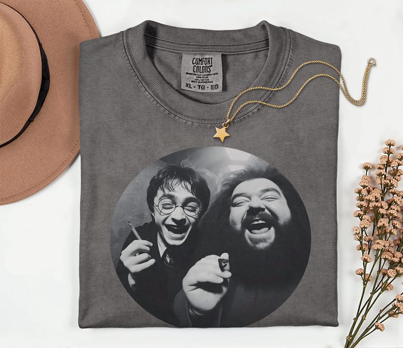 a gray t-shirt with a black and white photo of two men, one with glasses and the other with a beard, both smiling and laughing. The t-shirt is placed on a white surface, with a brown hat and some flowers nearby.