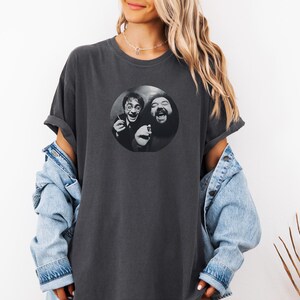 A woman wearing a gray t-shirt with a graphic of two men smiling and laughing, along with a denim jacket and a plant in the background.