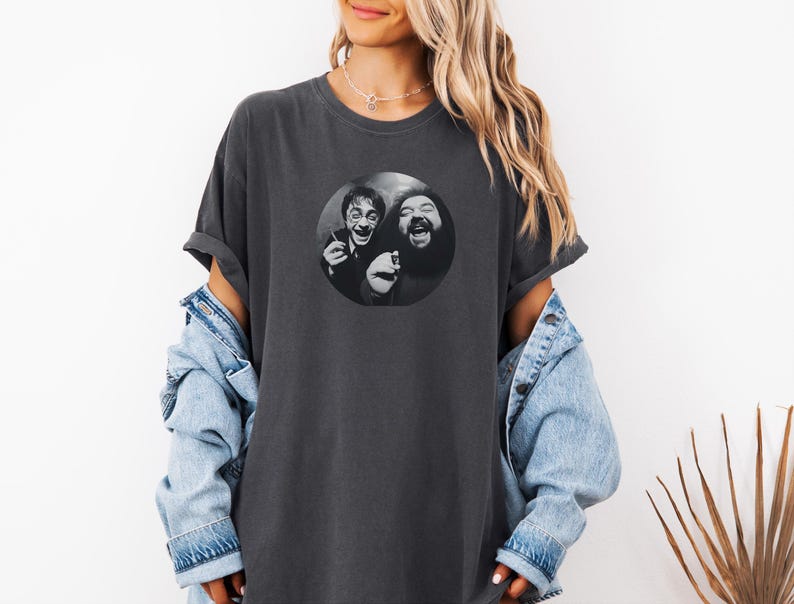 A woman wearing a gray t-shirt with a graphic of two men smiling and laughing, along with a denim jacket and a plant in the background.