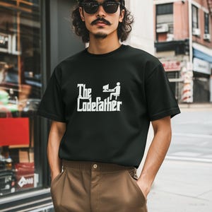 May include: A black short-sleeved t-shirt featuring a white graphic print on the chest. The graphic displays the text "The Codefather" in a stylized font, accompanied by a small white icon of a person sitting at a computer desk.