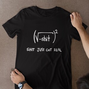 May include: A black short-sleeved t-shirt featuring a white graphic print of a mathematical equation with a square root of negative shit squared, followed by the text SHIT JUST GOT REAL printed in a hand-drawn style font on the chest.