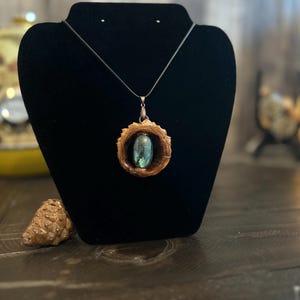 May include: A handcrafted pendant necklace featuring a natural wood frame with a central, iridescent, oval-shaped stone. The pendant hangs from a thin, dark cord, displayed on a black velvet jewelry stand. A pinecone rests on the wooden surface.