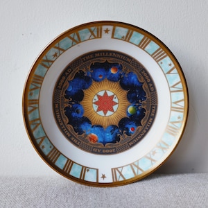 May include: A white decorative plate with a gold rim and a celestial design. The centre features a sunburst with planets and the text "THE MILLENNIUM 2000 AD". The outer rim has Roman numerals and a light blue border.