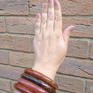 Puede incluir: Un conjunto de cuatro brazaletes lisos y redondeados en varios tonos de marrón, ámbar y naranja rojizo. Las pulseras presentan un acabado pulido y brillante con patrones veteados, apiladas juntas para mostrar sus distintos colores.