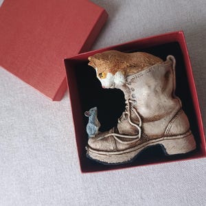 May include: A whimsical figurine of a cat peeking out of a weathered boot, with a small mouse perched on the boot's side. The figurine is presented in a red gift box with a black interior, perfect for a cat lover.