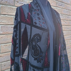 Puede incluir: Un chal gris con un patrón geométrico y diseños de cachemira. El chal presenta detalles de terciopelo burdeos y ribete de cuentas negro. La prenda está drapeada sobre un maniquí, mostrando su diseño fluido y sus intrincados detalles.