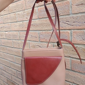 Könnte beinhalten: Eine hellbeige Umhängetasche aus Leder mit einer asymmetrischen, dunkelroten Fronttasche mit Klappe und einem passenden, verstellbaren Schulterriemen. Die Tasche hat eine glatte Oberfläche und ein kleines geprägtes Logo unten rechts.