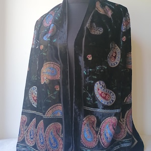Op de afbeelding: Een zwarte fluwelen sjaal met een paisley patroon in rood, blauw en goud. De sjaal is over een mannequin gedrapeerd, wat het gedetailleerde ontwerp en de zachte textuur benadrukt. De randen zijn afgewerkt met een donkerdere rand.